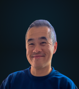 A man with short, neatly combed hair is smiling warmly at the camera. He is wearing a dark blue shirt, and the background is a dark, solid color.