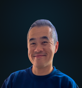 A man with short, neatly combed hair is smiling warmly at the camera. He is wearing a dark blue shirt, and the background is a dark, solid color.