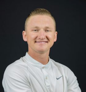 A man with short blond hair wearing a white Nike polo shirt stands with arms crossed, smiling confidently against a plain black background.