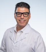 A man wearing black glasses and a white lab coat smiles at the camera with his hands in his pockets, standing against a plain light background.