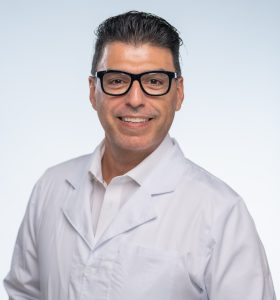 A man wearing black glasses and a white lab coat smiles at the camera with his hands in his pockets, standing against a plain light background.