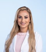 A woman with long blonde hair wearing a light pink dress and a white lab coat smiles while standing against a plain light blue background.