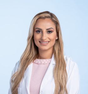 A woman with long blonde hair wearing a light pink dress and a white lab coat smiles while standing against a plain light blue background.