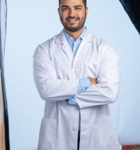 A man with dark hair and a beard, wearing a white lab coat over a light blue shirt, stands smiling with arms crossed in a studio setting.