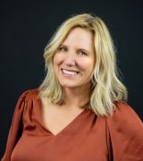 A woman with shoulder-length blonde hair, wearing a rust-colored blouse, smiles in front of a solid black background.