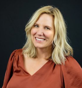 A woman with shoulder-length blonde hair, wearing a rust-colored blouse, smiles in front of a solid black background.