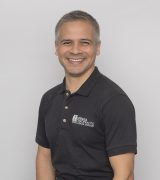A man with short gray hair wearing a black polo shirt and dark pants sits on a stool, smiling at the camera against a plain light gray background.