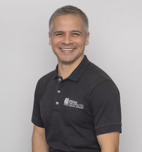 A man with short gray hair wearing a black polo shirt and dark pants sits on a stool, smiling at the camera against a plain light gray background.