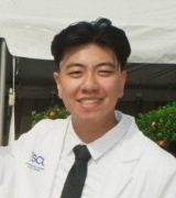 A young person with short black hair smiles at the camera while wearing a white lab coat with an ISCL logo and a black tie, standing outdoors in front of greenery and a white tent.