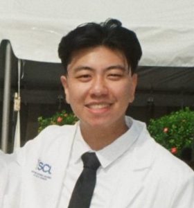 A young person with short black hair smiles at the camera while wearing a white lab coat with an ISCL logo and a black tie, standing outdoors in front of greenery and a white tent.