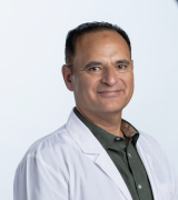 A man wearing a white lab coat over a dark green button-up shirt stands against a plain, light background, smiling slightly and looking at the camera.
