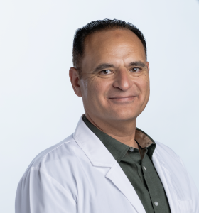 A man wearing a white lab coat over a dark green button-up shirt stands against a plain, light background, smiling slightly and looking at the camera.