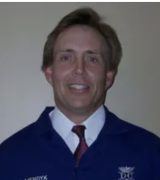 A man in a blue jacket embroidered with Dr. Mendyk stands against a plain background, wearing a white shirt and red tie, smiling at the camera.