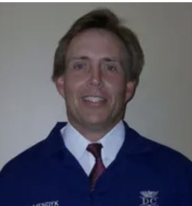 A man in a blue jacket embroidered with Dr. Mendyk stands against a plain background, wearing a white shirt and red tie, smiling at the camera.
