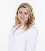 A woman with wavy blonde hair wearing a white lab coat stands smiling with her hands in her pockets against a plain white background.