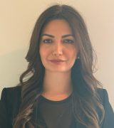 A woman with long, wavy dark hair is standing against a plain light-colored wall. She is wearing a black blazer over a black top and is looking directly at the camera with a neutral expression.