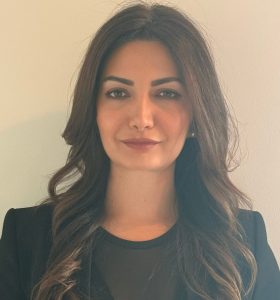 A woman with long, wavy dark hair is standing against a plain light-colored wall. She is wearing a black blazer over a black top and is looking directly at the camera with a neutral expression.