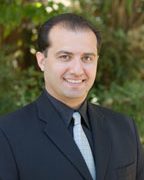 A man with short dark hair, wearing a black suit, light gray tie, and white shirt, smiles at the camera. Greenery and trees are blurred in the background.