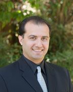 A man with short dark hair, wearing a black suit, light gray tie, and white shirt, smiles at the camera. Greenery and trees are blurred in the background.