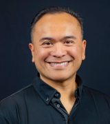 A man with short dark hair, wearing a black button-up shirt, smiles while standing against a plain black background.