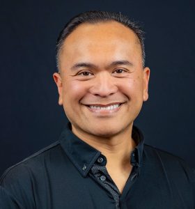 A man with short dark hair, wearing a black button-up shirt, smiles while standing against a plain black background.