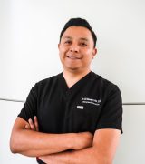 A man in black medical scrubs stands against a white background with arms crossed, smiling at the camera. His shirt reads Dr. Gil Navarrete, D.D.S. Orthodontist/Prosthodontist.