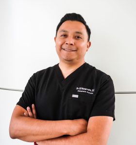 A man in black medical scrubs stands against a white background with arms crossed, smiling at the camera. His shirt reads Dr. Gil Navarrete, D.D.S. Orthodontist/Prosthodontist.