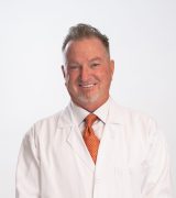 A man with gray hair and a goatee, wearing a white lab coat and an orange tie, stands smiling with hands clasped in front of a plain light background.
