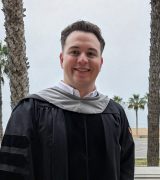 A person in a black graduation gown stands smiling on a balcony with palm trees and a beach in the background.