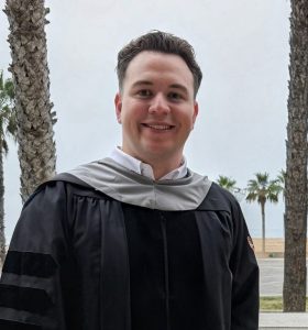 A person in a black graduation gown stands smiling on a balcony with palm trees and a beach in the background.