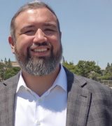 A man with a short gray beard and short hair, wearing a white dress shirt and a gray plaid blazer, stands outdoors on a sunny day with trees and a clear blue sky in the background.