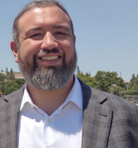 A man with a short gray beard and short hair, wearing a white dress shirt and a gray plaid blazer, stands outdoors on a sunny day with trees and a clear blue sky in the background.