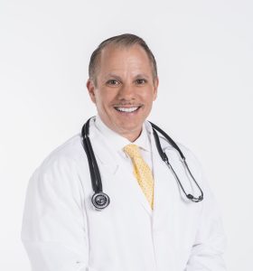 A smiling male doctor wearing a white lab coat, yellow tie, and stethoscope around his neck stands against a white background.