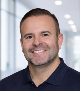 Smiling man with short brown hair and neatly trimmed beard, wearing a dark blue polo shirt, standing indoors with a blurred bright background.