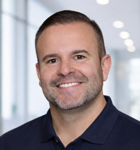 Smiling man with short brown hair and neatly trimmed beard, wearing a dark blue polo shirt, standing indoors with a blurred bright background.