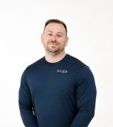 A man with short hair and a trimmed beard wears a navy blue long-sleeve shirt with SADA written on it, standing against a plain white background and smiling slightly.