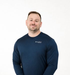 A man with short hair and a trimmed beard wears a navy blue long-sleeve shirt with SADA written on it, standing against a plain white background and smiling slightly.