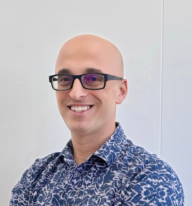 A smiling person with glasses, wearing a blue and white patterned shirt, stands against a plain white background.